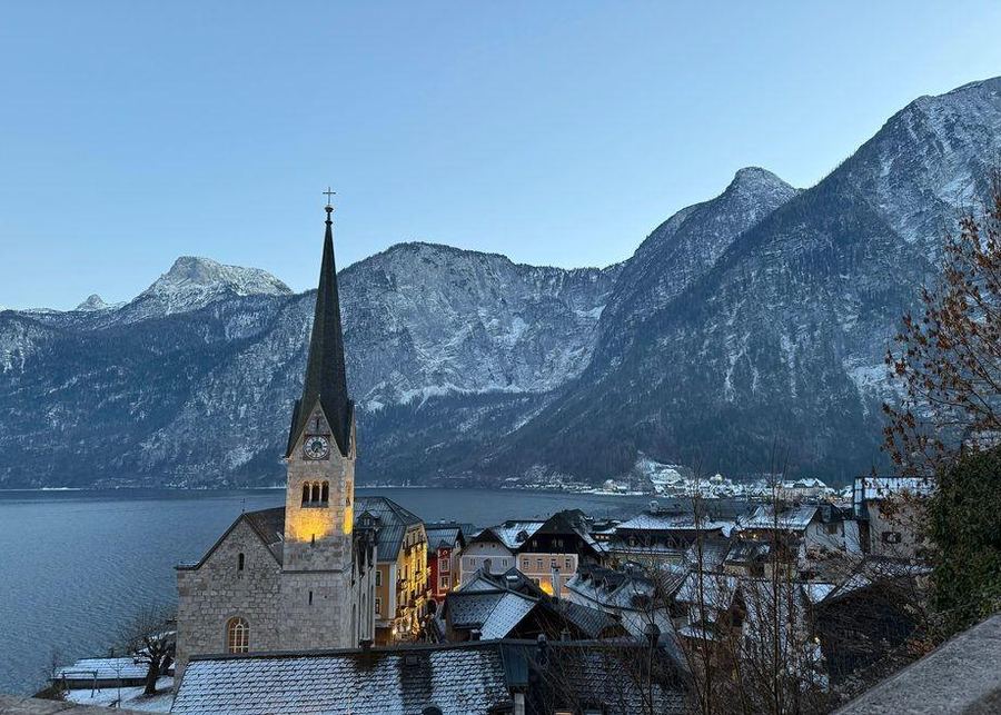 A lakeside village of snow-dusted roofs below steep snowy mountains, with a lit church spire in the foreground at dusk