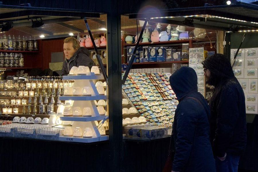 A lit market hut selling ceramic tea-light houses, fridge magnets and painted postcards, with two hooded shoppers looking in from the dark