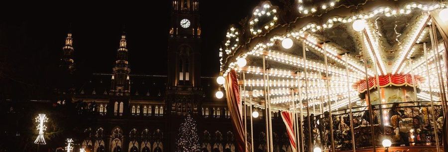 A carousel covered in light bulbs turning at night in front of Vienna's floodlit neo-gothic City Hall