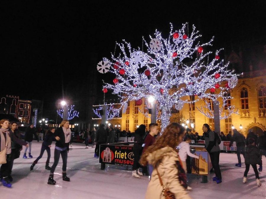 Skaters on an outdoor ice rink at night around a tree of white lights hung with red baubles and paper snowflakes, in front of a floodlit gothic building