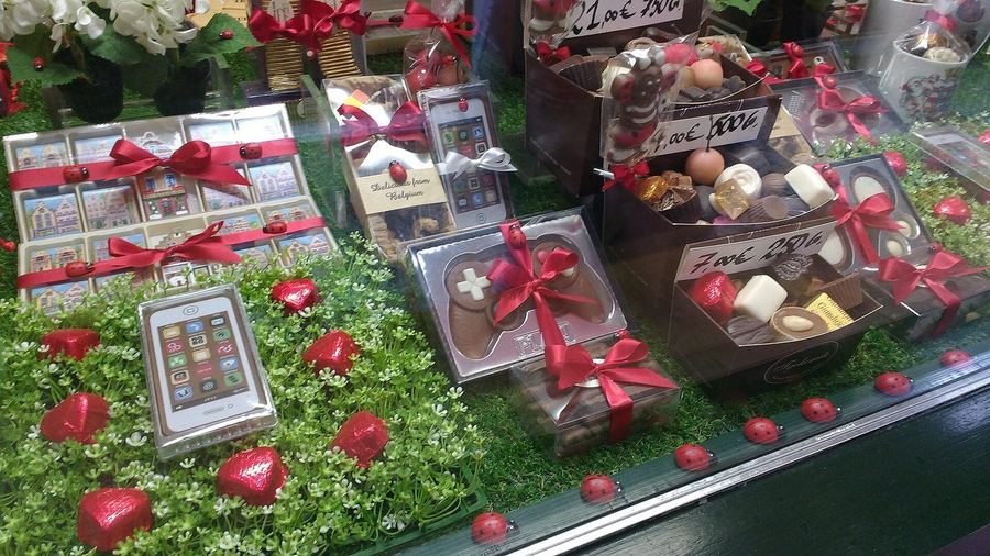 A chocolate shop window packed with ribboned boxes of pralines and moulded chocolates, each box priced by weight in euros