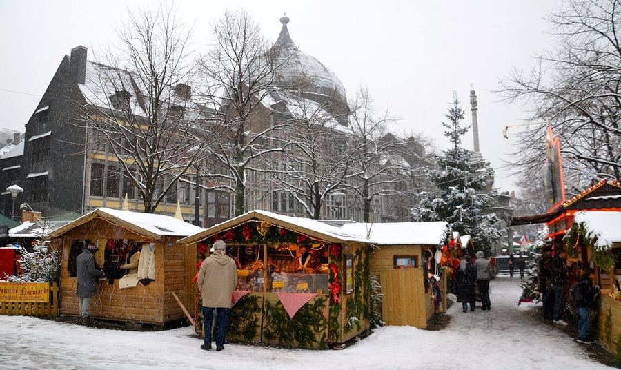 Wooden market chalets under falling snow, with bare trees, a lit stall of nuts and a domed building behind