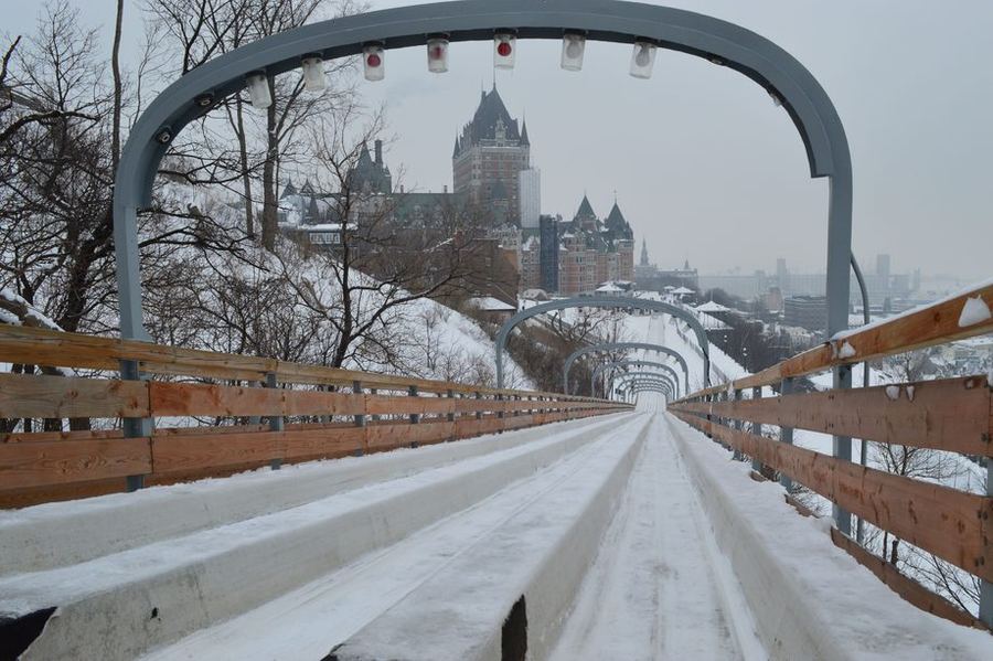 A snow-packed toboggan run under metal arches on Quebec's Dufferin Terrace, with the turreted Chateau Frontenac above it