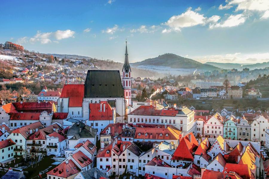 Red-tiled roofs of a small Czech town in winter sunshine, with a tall white church spire and snow on the hills behind