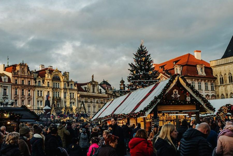 A dense crowd at a Christmas market in a baroque square at dusk, with a tall decorated tree and a garlanded wooden market hall behind