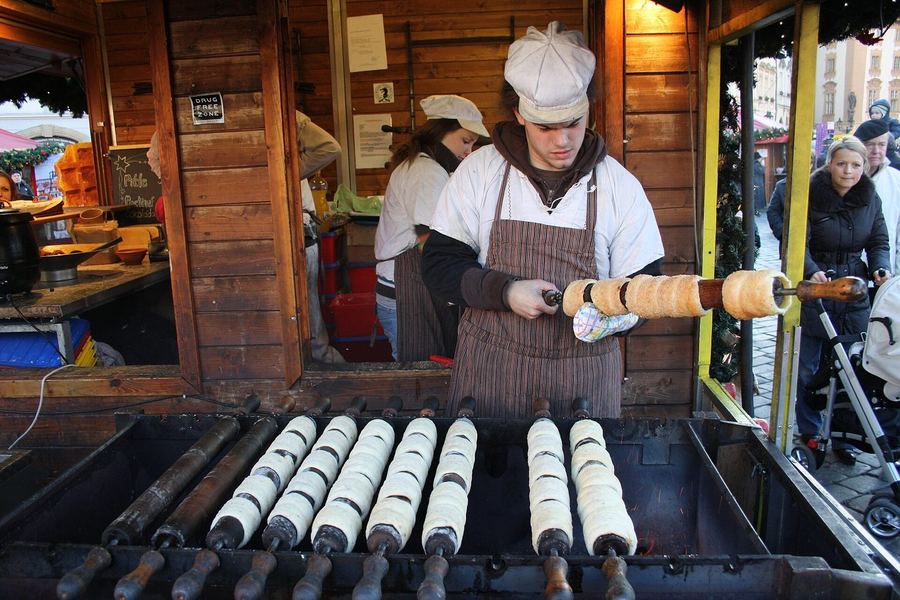 A cook in an apron turning dough-wrapped spits of trdelník over a charcoal trough at a wooden market stall