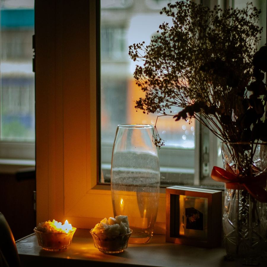 Candles burning in two small glass bowls on a windowsill at dusk, beside dried flowers and a framed photograph