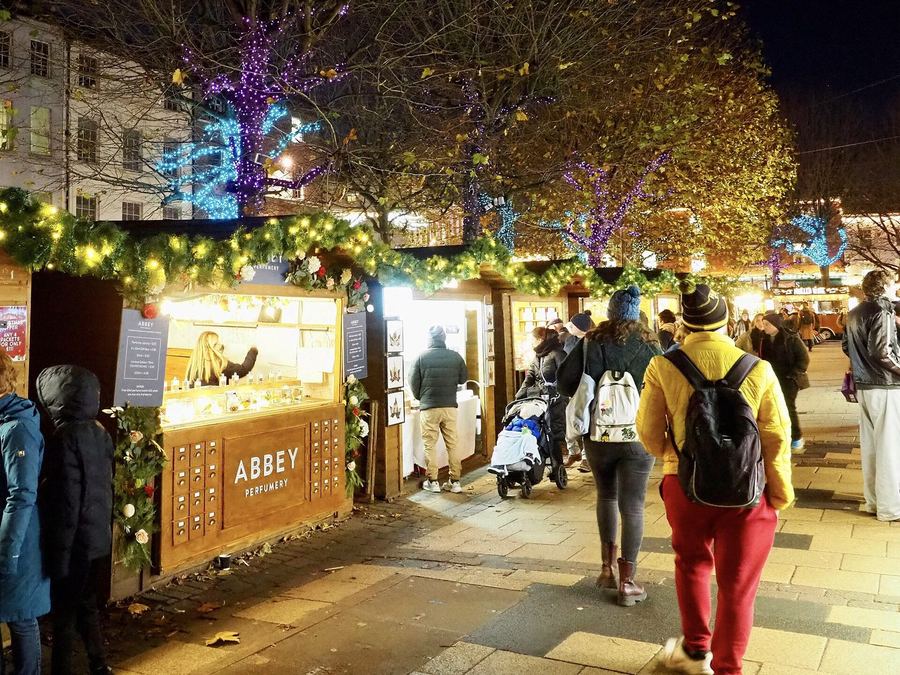 Lit wooden market chalets hung with garlands at night, shoppers in winter hats walking past under trees strung with lights