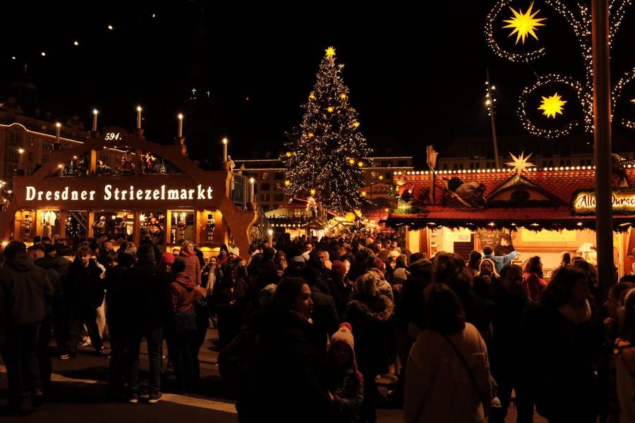 A dense night crowd under a lit tree beside a wooden arch reading Dresdner Striezelmarkt