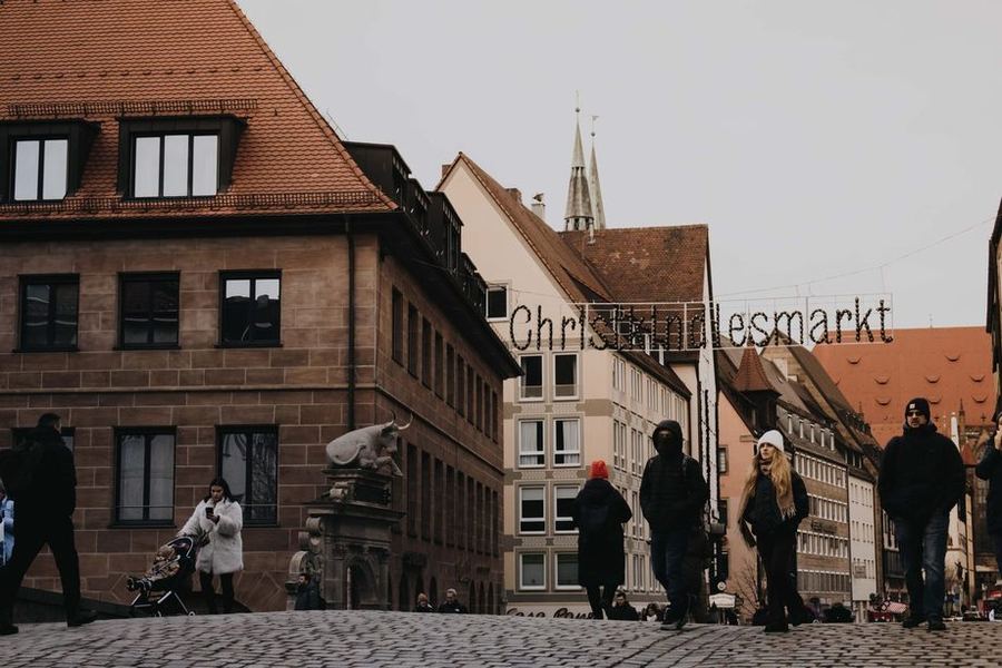 A lit Christkindlesmarkt sign strung across a cobbled Nuremberg street on a grey day, with people walking up towards it