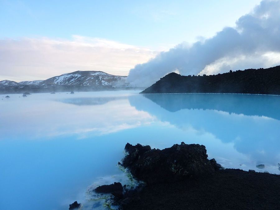 Steam rising off pale blue water between black lava rocks, with snow-streaked hills behind