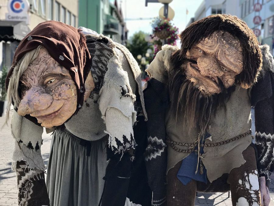 Two giant troll figures with warty faces and ragged woollen clothes, leaning over a sunlit pedestrian shopping street