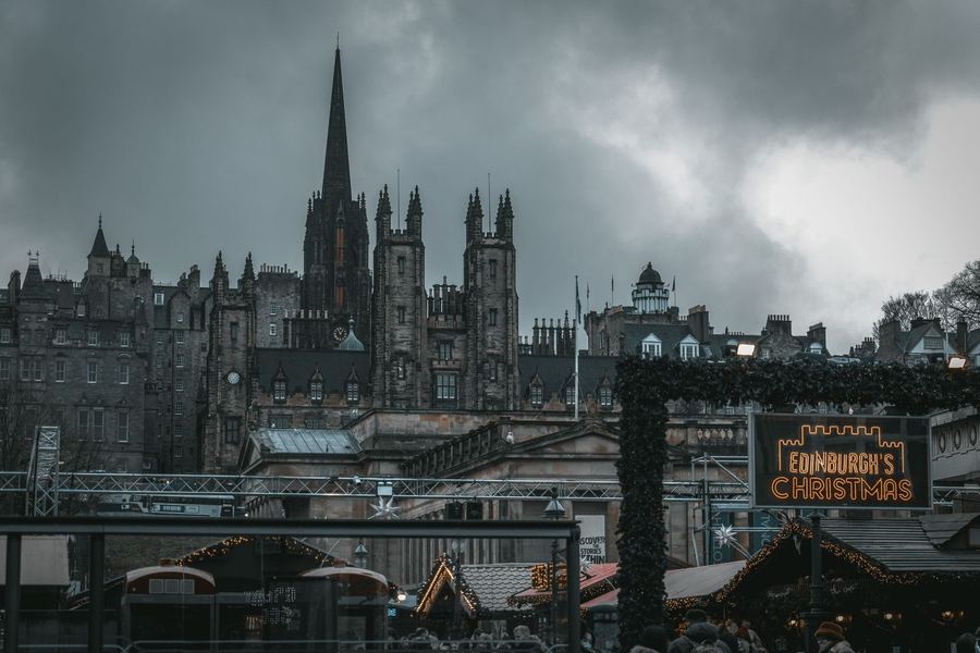 Lit market huts and a neon Edinburgh's Christmas sign below the spires and gothic towers of the Old Town under a heavy grey sky