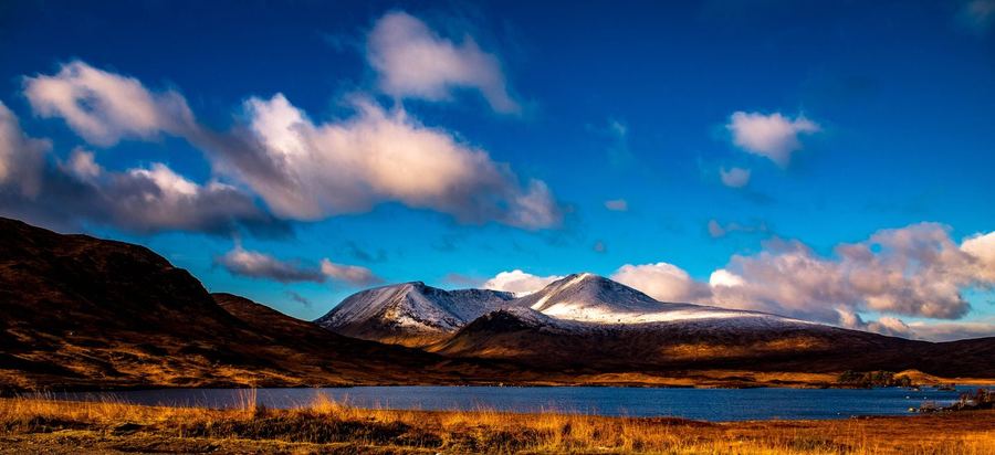 Snow-topped mountains above a loch and pale winter grass under a bright blue sky with white cloud