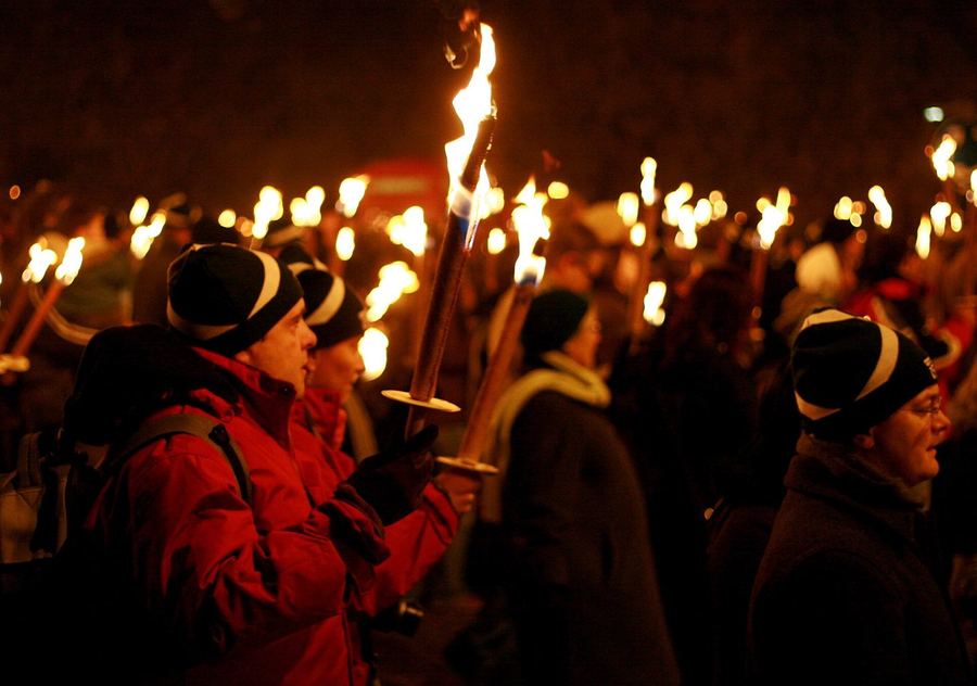 A night-time crowd in woolly hats and winter coats carrying burning torches along a street