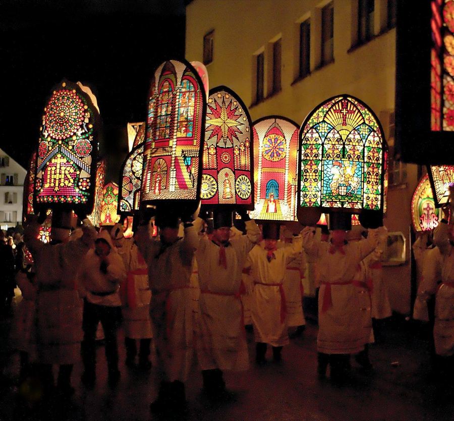 Men in white robes and red sashes carrying tall mitre-shaped headdresses lit from inside like stained glass, through a dark street at night