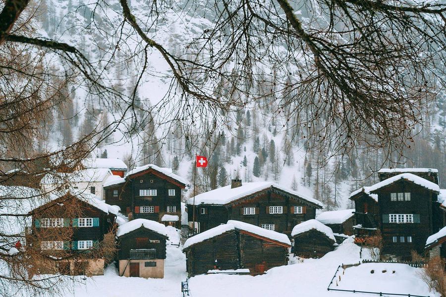 Dark timber chalets under deep snow on a wooded mountainside, with a Swiss flag flying among them