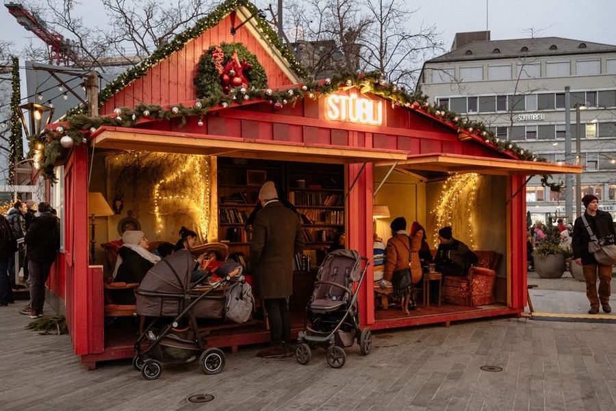 A red wooden market chalet signed Stübli, open at the front with armchairs, bookshelves and fairy lights inside, and prams parked outside at dusk