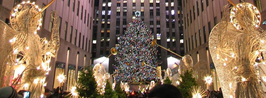 A huge lit Christmas tree in front of a skyscraper, framed by wire-frame angel sculptures blowing trumpets